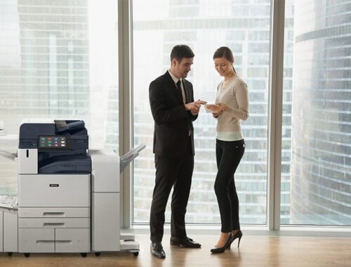 Deux personnes en tenue professionnelle dans un bureau discutent de documents à côté d'une imprimante multifonction Xerox® AltaLink® série B8200